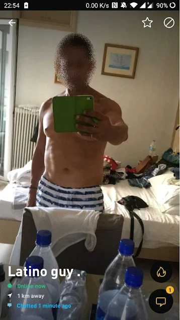 Screen shot of a Grindr profile, showing a guy taking a selfie of himself in his very messy hotel room