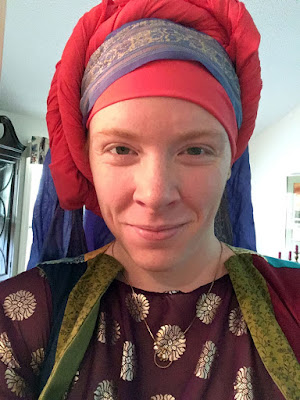 A selfie of a white woman smiling mostly with her eyes, wearing a tall, twisted coral-pink turban with a band of purple-and-gold brocade over the crown of her head, and a translucent purple veil falling from the back of the turban. She has a red-violet boat-necked shirt with gold stamped flower roundels all over, and a patchwork stripe vest with green, red, and blue strips. Her thin gold necklace almost disappears against the shirt, but the gold hoop pendant shows pretty clearly.