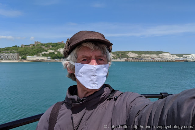 John Latter on the New Marina Pier, Dover Western Docks Revival (DWDR) area of the Port of Dover, Kent, England, wearing a homemade Coronavirus personal protection equipment face mask. Dover Castle's Saxon St Mary-in-Castro church and Roman Pharos on the White Cliffs in background.