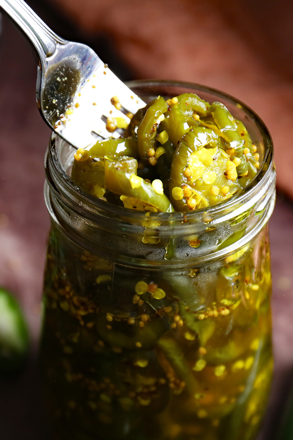 A close-up view of the top of a jar filled with enticing cowboy candy. A fork is skillfully digging into the jar, gently lifting up the mouthwatering candied jalapenos, tempting the taste buds with their sweet and spicy allure.
