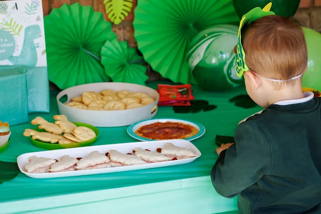 A boy wearing a dinosaur mask eating dinosaur themed food which is on a decorated table