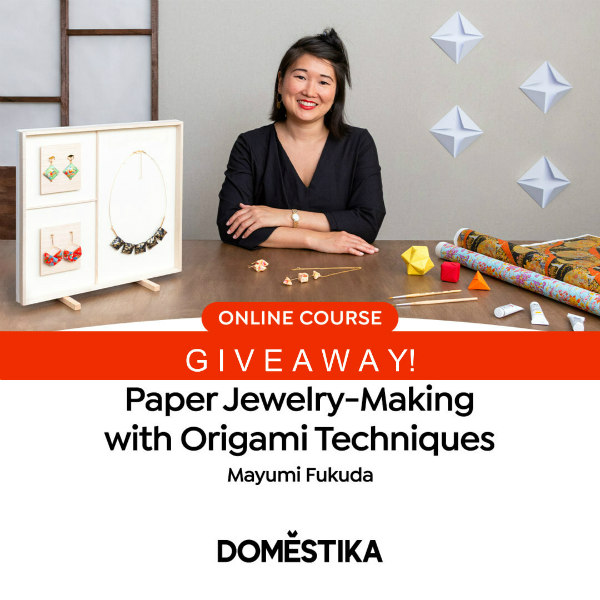smiling woman seated at table on which origami jewelry and paper supplies are displayed