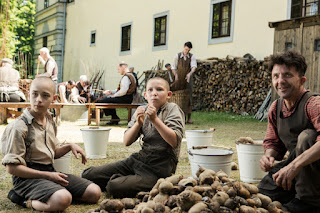 Two boys with shaved heads(Niklas Post and Ivo Pietzcker) and a man (David Bennent) sit outdoors by a pile of potatoes looking amused and shocked. Behind them, men are making baskets.  Courtesy of StudioCanal.
