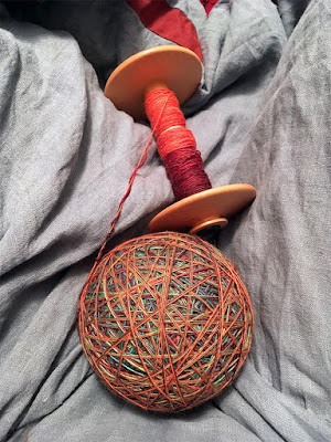 A large, many-colored ball of wrapped thread with crisscrossing strands in red, orange, blue, green, and yellow, attached to a wooden bobbin with a single layer of thread fading from orange to deep pink-red, held in the photographer's blue-grey skirted lap.