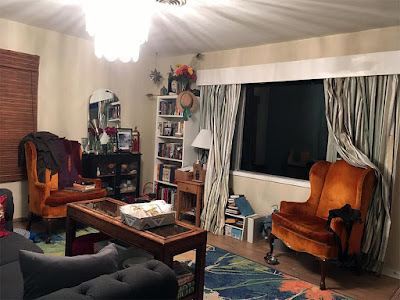 A living room with a number of bookshelves, two orange velvet armchairs, and a small cabinet full of yarn, lit by an extremely bright chandelier.