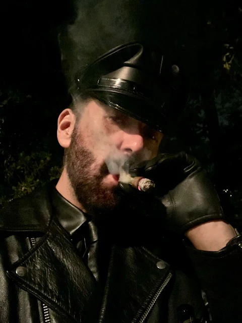 Smoking cigar wearing full black leather uniform including biker jacket and Muir cap puffing a gar chest up