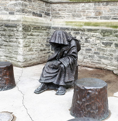 bronze sculpture of figure in sandals and a hooded cloak, seated in front of an old stone wall