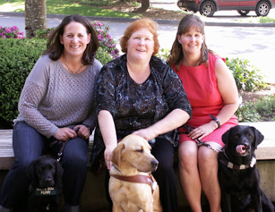 Megan, Laura Ann and Cathy sit smiling on a bench with their dogs