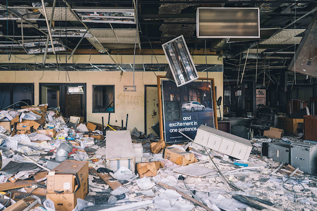 Chaos reigns in the interior of a devastated showroom, with collapsed ceilings, scattered documents, and an overturned promotional display.