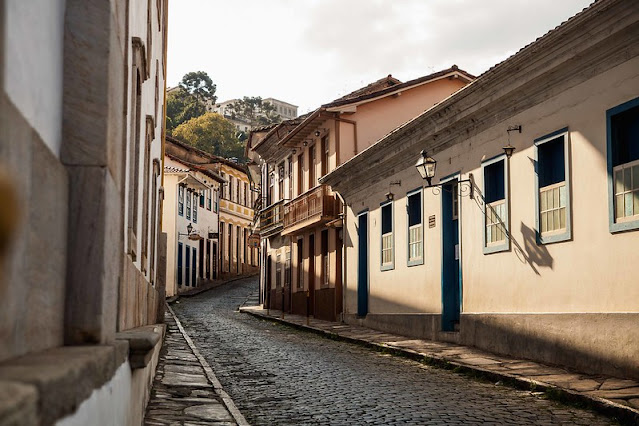 A fotografia mostra uma rua no centro histórico de Ouro Preto. O sol ilumina as casas do lado esquerdo da rua. Não há pedestres nem trânsito.