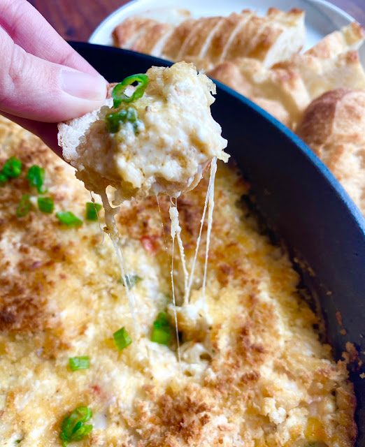 Hand holding a cheesy section of baked seafood dip with long strands of cheese pulling from the skillet.