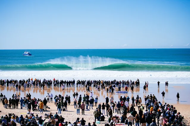 Paisagem de Peniche, com uma onda com tubo grande no fundo e muitas pessoas na faixa de areia da praia, formando três colunas espaçadas de pessoas