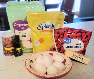 ingredients for the recipe, packages shown. 1 bowl with uncracked eggs, 1 bag of coconut flour, 1 bag of Splenda sugar blend, 1 stick of butter, 1 bottle of vanilla extract, 1 jar of baking powder, 1 package of frozen raspberries