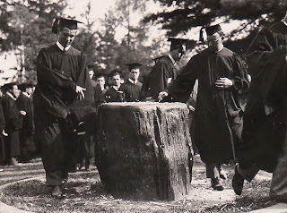 A group of figures in caps and gowns. In the foreground, two men are touching a tree stump.