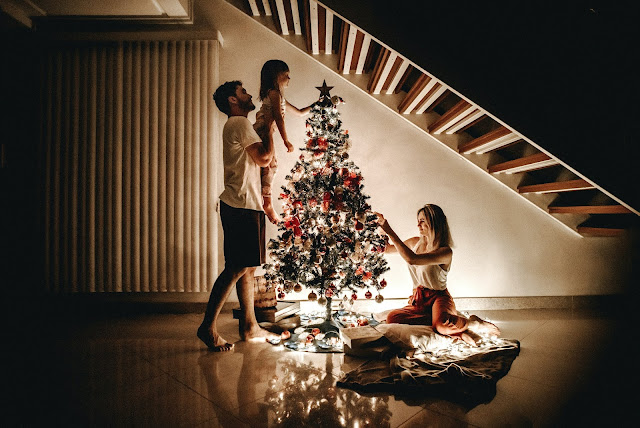 Mujer decorando árbol de navidad bajo una escalera. Hombre levantando a niña decorando el mismo árbol de navidad