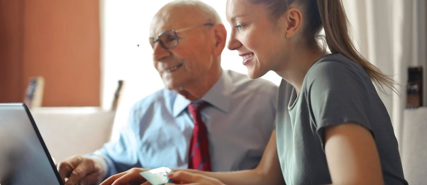 An elderly person sat next to a younger person who is holding a debit/credit card. They're both looking at a laptop screen and the younger person is helping input financial details.