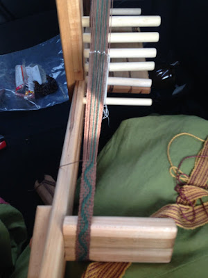 A wooden inkle loom with a brown and green warp and an undulating green stripe in progress, resting partly on the photographer's green-skirted and rose-and-gold-belted lap.