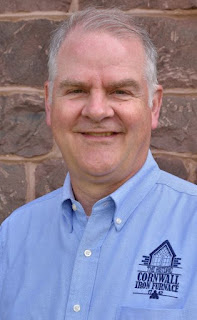 White man with graying hair stands in front of a brown stone wall. He's wearing a blue button-down shirt with the words "Cornwall Iron Furnace" in the right of the photo.