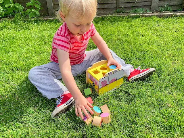 Toddler reaching out for coloured blocks to put in yellow wooden peppa pig campervan shape sorter