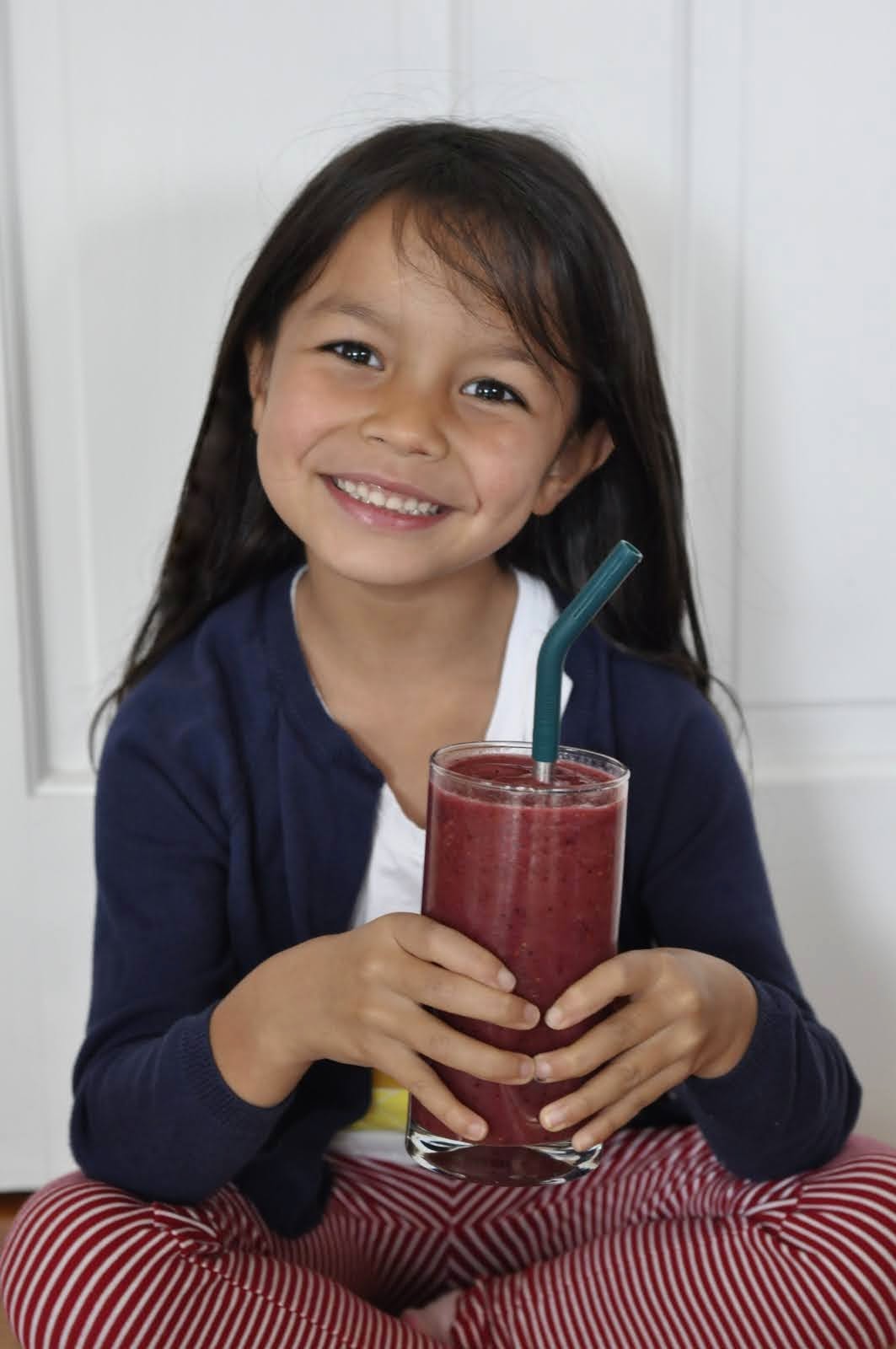 Young child with long brown hair holds a glass with a Simple Mixed Berry Smoothie in it. The glass also has a metal straw to make sipping a lot easier.