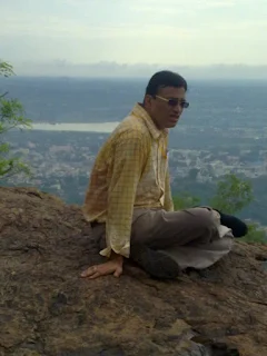 Kasturi Rangan Varadan seated on a rocky hilltop with a panoramic view of a town and serene surroundings in the background.