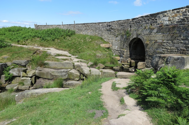 Looking at a stone bridge from below. An arch allows the brook to flow beneath the road from the moors above down into the valley.