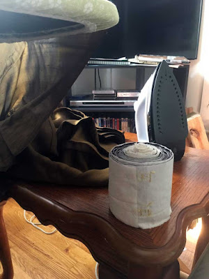 A short roll of fabric, white on the outside with brown peeping out at the edges, on a wooden table with an iron and a pile of green-gold fabric, pooling where it falls off the pale-green floral ironing board at topmost left of the photo.