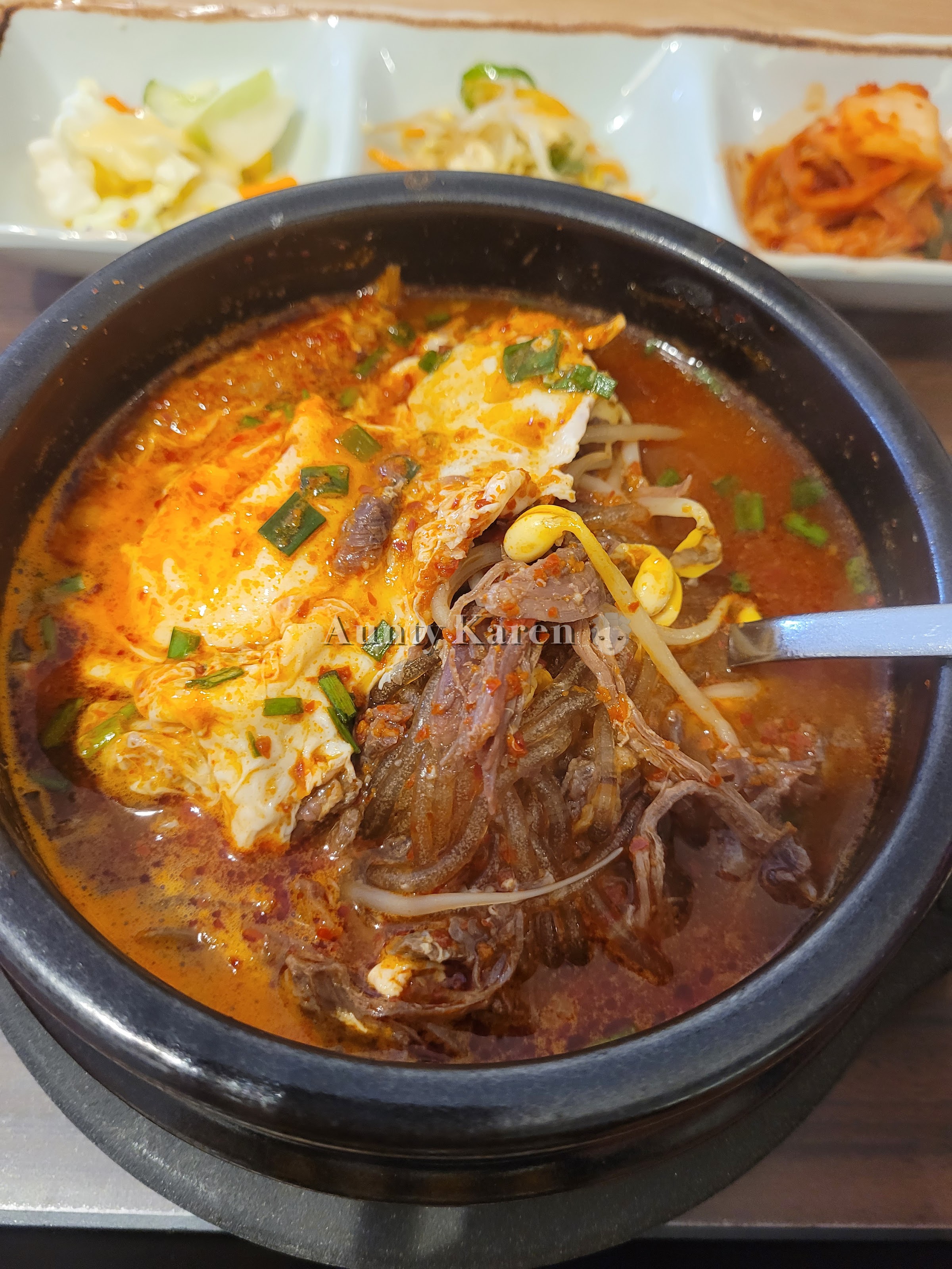 CLose-up of a bowl of Korean spicy beef soup with a trio of side dishes in the background