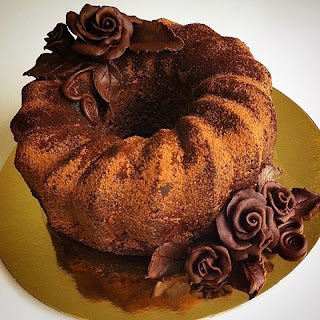 A foto mostra um bolo de chocolate redondo, alto em forma de gomos, salpicado por cacau em pó. Sobreposto à esquerda, na superfície, um arranjo em formato de uma rosa cercada por folhas também de chocolate. Na diagonal, sobre a base de papelão dourado e redondo, outro arranjo floral maior, no mesmo estilo com três rosas.