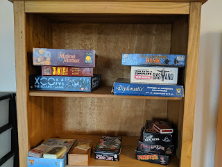 Two shelves of a bookcase with a few games on, including "XCOM The Board Game", "Junta!", a French edition of "Diplomacy", "Medieval Merchant", "Zombies!!!" and a box of poker chips.