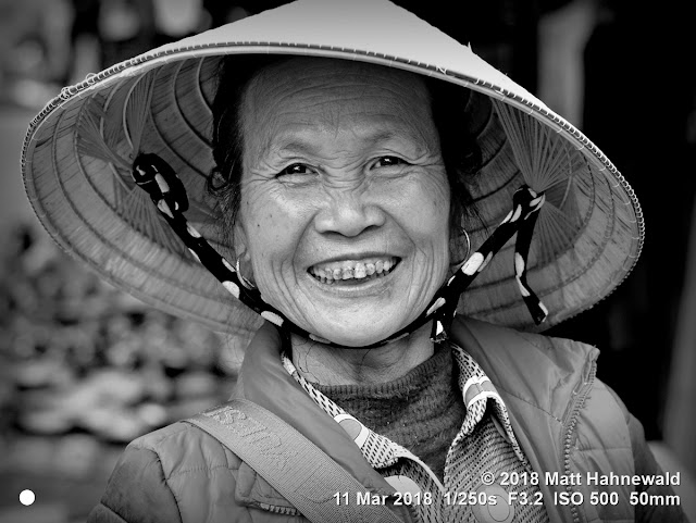 matt hahnewald photography; facing the world; basic; character; head; face; eyes; catchlights; smiling eyes; teeth; wrinkles; expression; looking at camera; headgear; headwear; hat; conical hat; sun hat; rice hat; asian hat; consent; emotion; concept; humanity; culture; tradition; enjoyment; optimism; sisterhood; traditional; cultural; market; bac ha; lao cai; northern; vietnam; asia; asian; vietnamese; individual; one person; female; elderly; woman; image; photo; physiognomy; nikon d3100; nikkor afs 50mm f1.8g; prime lens; 50mm; 4x3; horizontal; street; portrait; closeup; headshot; full face view; outdoor; mono; black and white; monochrome; greyscale; vignette; posing; smiling; happy; joyous