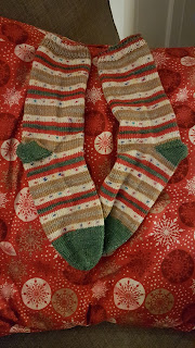 A pair of finished hand knit socks. The have green heels and toes and stripe in red, green, gold, silver and an amazing white with little coloured dots to represent fairy lights. They are lying on a christmas pillow.