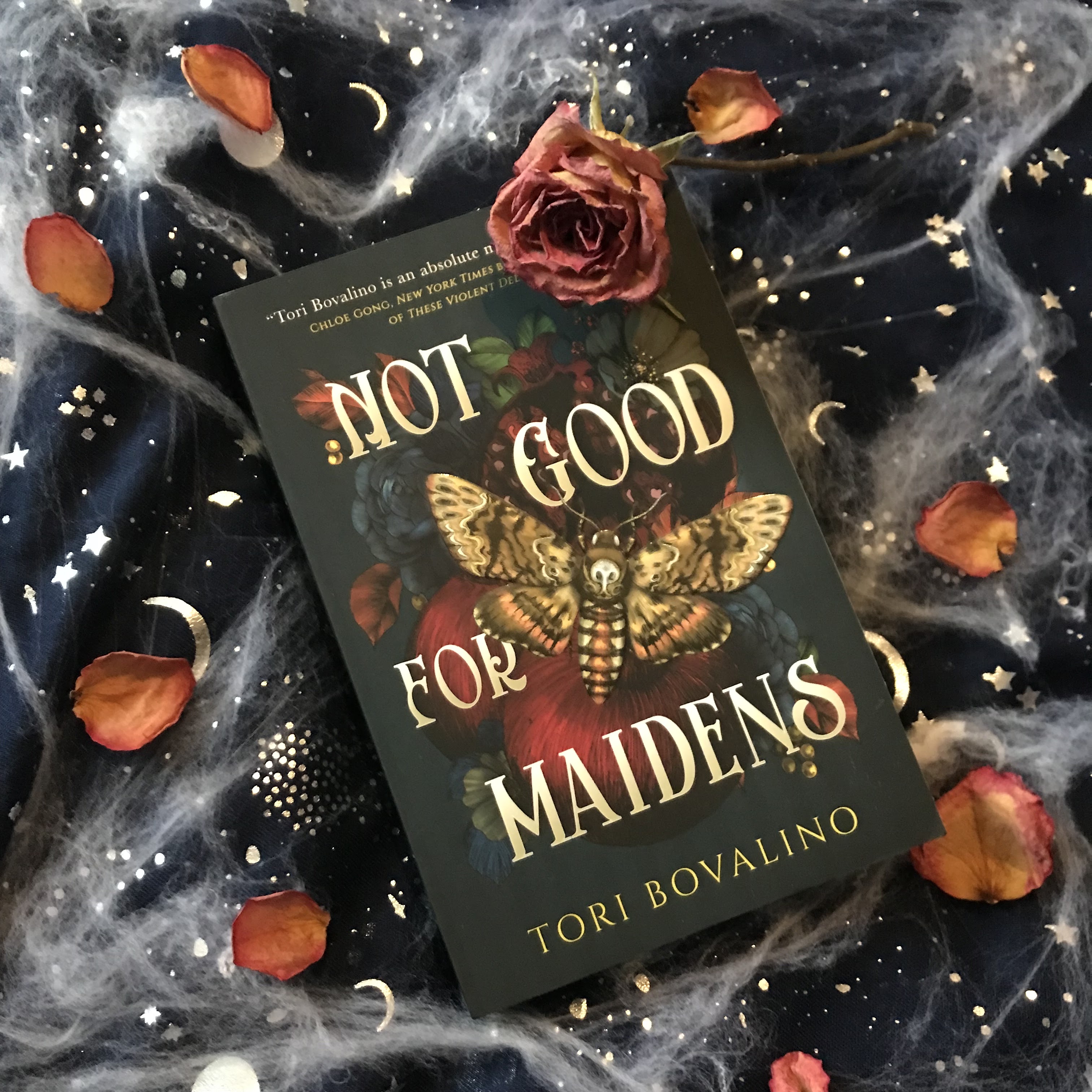 Not Good for Maidens by Tori Bovalino is on a deep blue scarf with metallic silver moons and stars, on a diagonal top left to bottom right. Around it are fake cobwebs. On the top right corner of the book is a dried rose, and rose Bryan’s are scattered around the book.