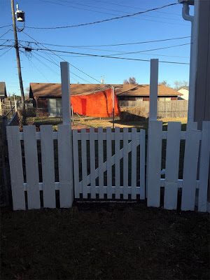 A white picket gate hanging between white-painted posts with short sections of fence on either side, strongly backlit by a sunny yard with a huge length of orange-gold fabric hanging on the clothesline.