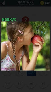 Девочка в саду делает надкус на яблоке, которое висит на дереве