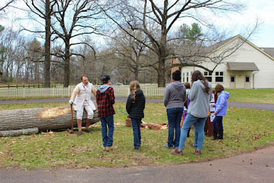 A white man in knee breeches and long waistcoat talks with a group of homeschool students. He is standing in front of a large fallen tree with some of the bark shaved off and is hold a long-handled tool to demonstrate.