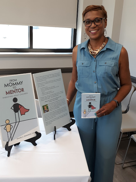 Pamela Taylor is standing to next to a table with her books, smiling. She is holding one of her books.