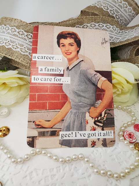 Postcard of 1950's housewife smiling in kitchen. She's in a blue dress with a white apron, has her hand on her hip and a dish towel glasped in her hand. Print reads " a career. . . a family to care for . . .gee! I've got it all!