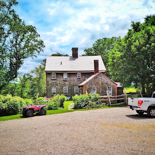 A late Georgian shingle-sided farmhouse across a gravel road surrounded by gardens