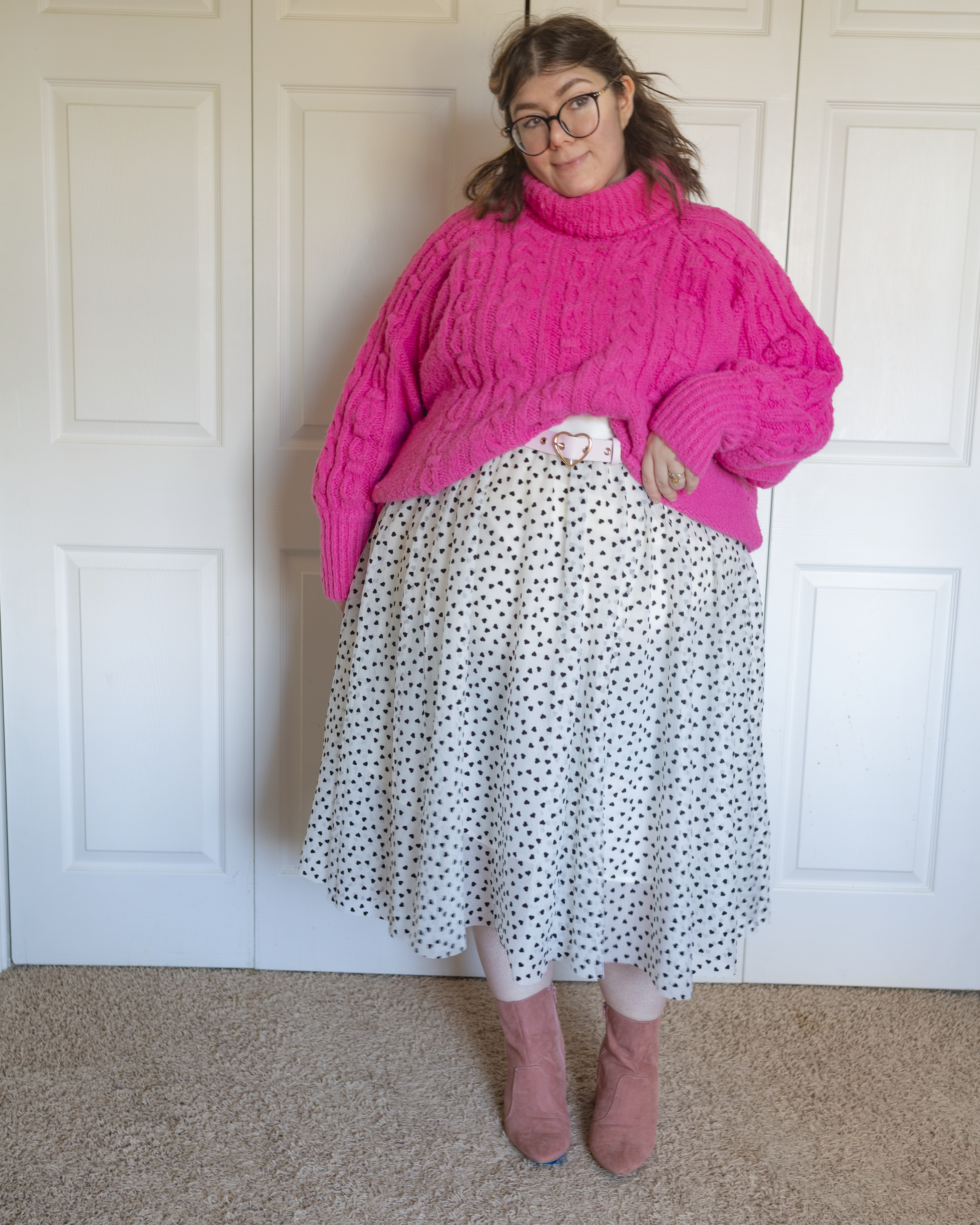 An outfit consisting of a hot pink cable knit turtleneck sweater half tucked into a white tulle midi skirt with heal hearts in a polka dot pattern and pastel pink midi boots.