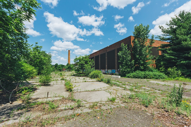 An expansive view of an overgrown concrete lot leading to an industrial brick building with graffiti, a tall smokestack in the distance, and a bright blue sky with scattered clouds overhead.