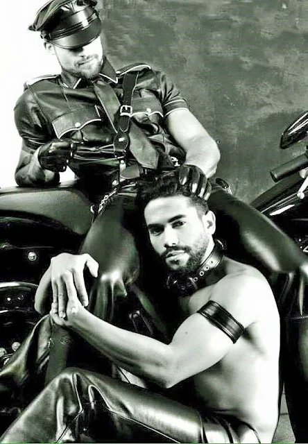 BW leather daddy sitting on motorcycle wearing gear running fingers through hair of slave kneeling below