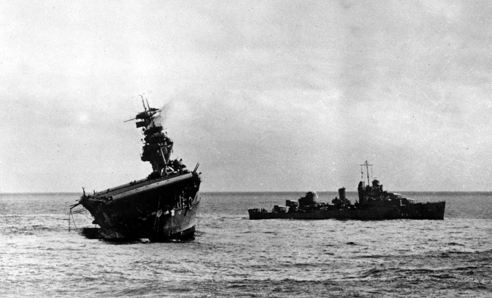 O USS Yorktown lista pesadamente ao porto depois de ser atingido por bombardeiros e torpedos japoneses na Batalha de Midway em 4 de junho de 1942. Um destróier fica à direita para ajudar enquanto uma tripulação de resgate no convés de vôo tenta consertar o porta-aviões .