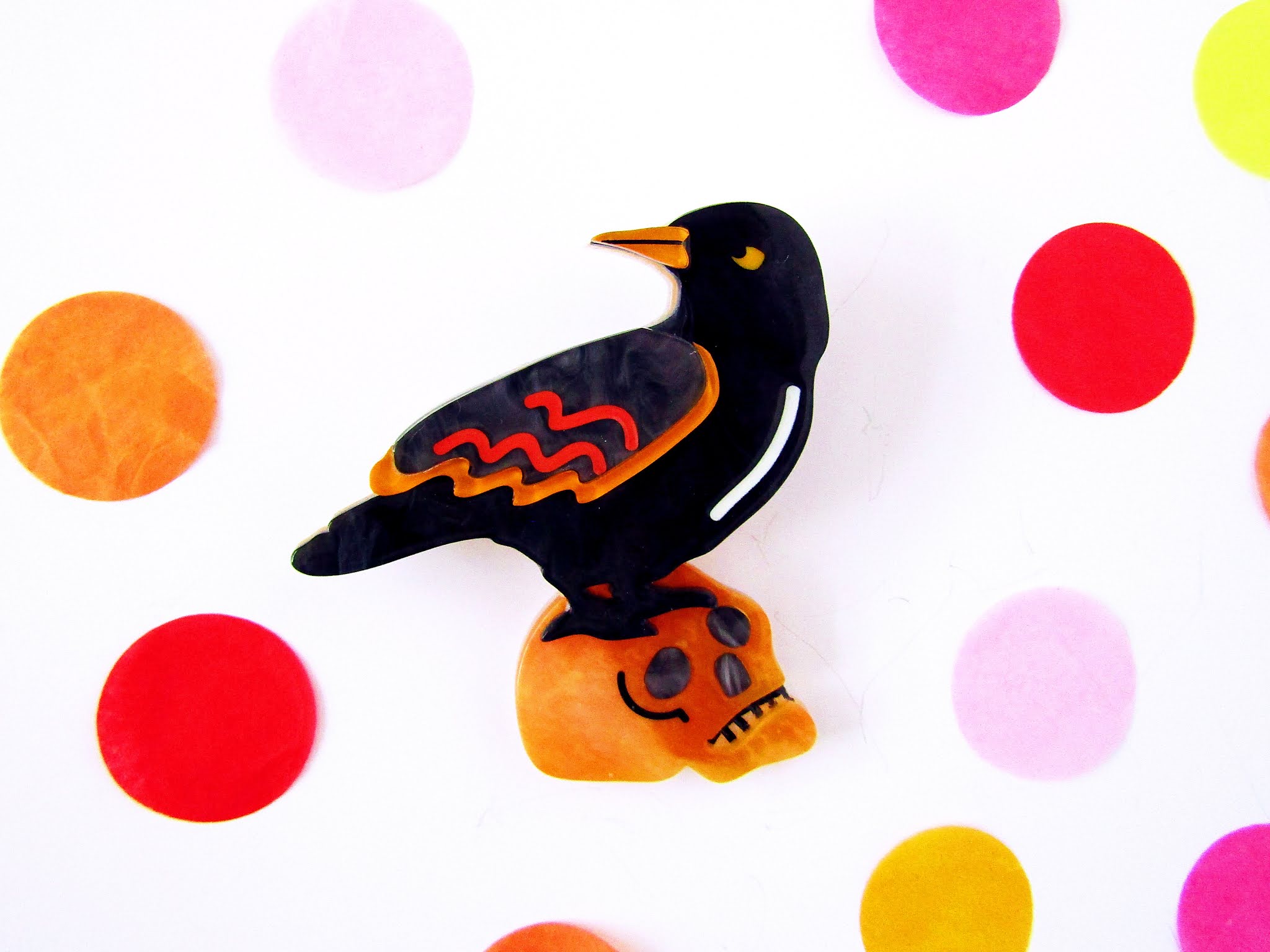 A photo of an Erstwilder black raven brooch on a white background surrounded by colourful confetti. The brooch features a black resin raven with orange markings on its wing perched on the top of an orange marbled resin skull.