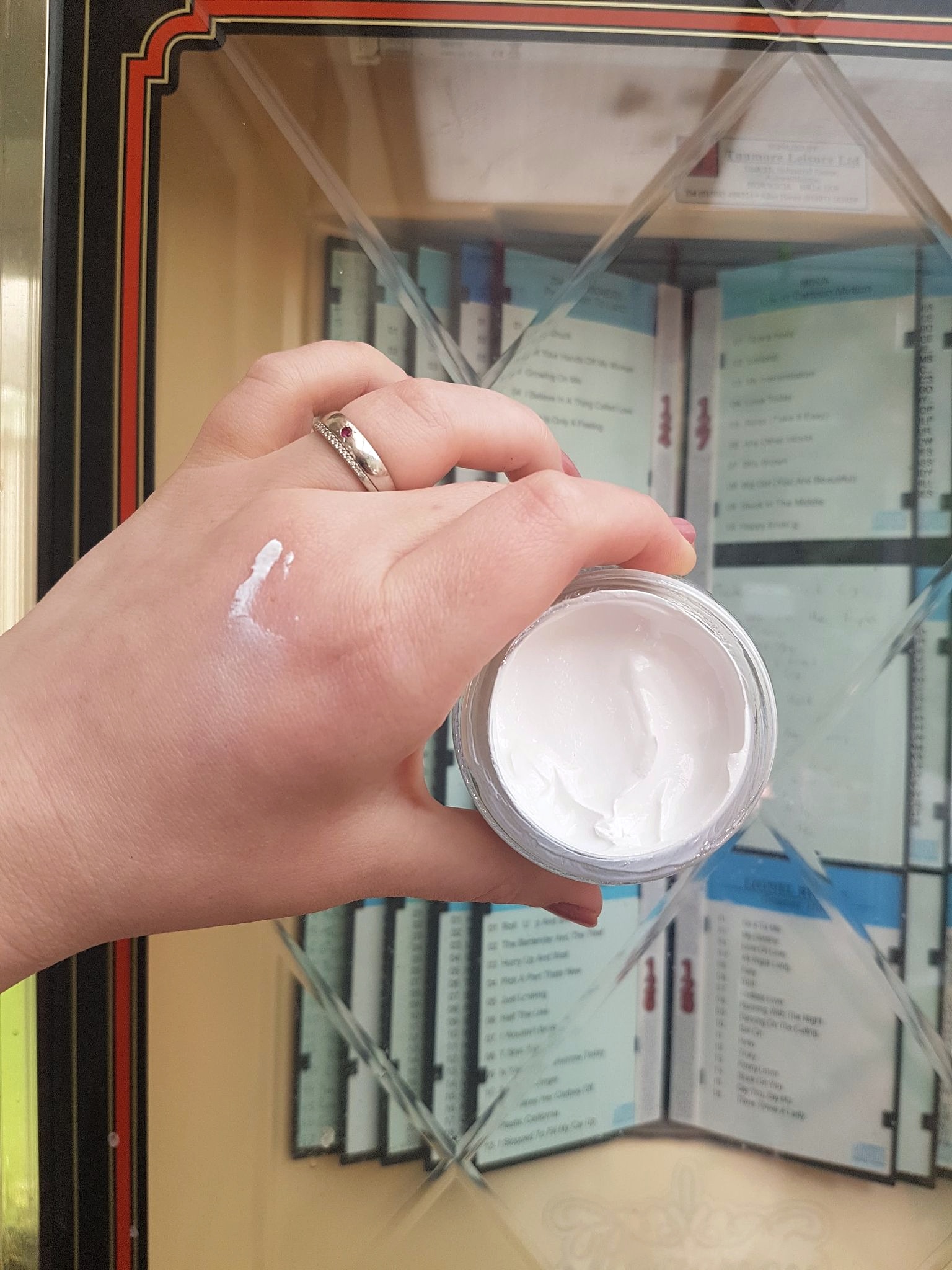 a glass jar filled with white reef safe sunscreen held up in front of a jukebox. a small amount is applied to the hand holding the jar