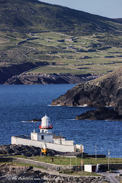 A white and red lighthouse by a dark blue ocean in front of green hills that are built throughout with low stone walls.