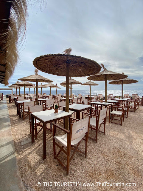 Rows and rows of empty brown wooden bistro tables with white table tops and wooden and white director chairs under straw sun umbrellas on a pebbled beach.