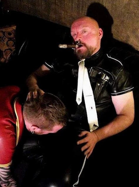 Leather master sitting superior above a kneeling sexy muscle slave giving head while he puffs a cigar like an kinky pimp for adults over 18 years.