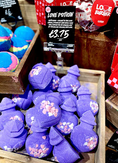 A wide angled photo of a large rectangular box containing a bunch of purple bottle shaped bath bombs on a bright background