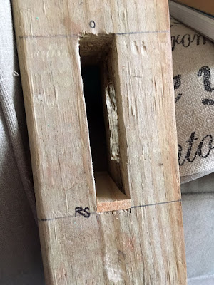 A mortise in the side of a 4-by-4, marked with faded pen lines and a small circle drawn directly above and "RS" at lower left. A lighter piece of wood is fitted into the base of the mortise; looking into the opening, a second opening on the adjacent face of the beam is also visible.